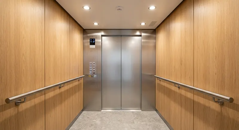 Modern elevator cab with light oak wall panels, a stainless steel handrail, and warm lighting.